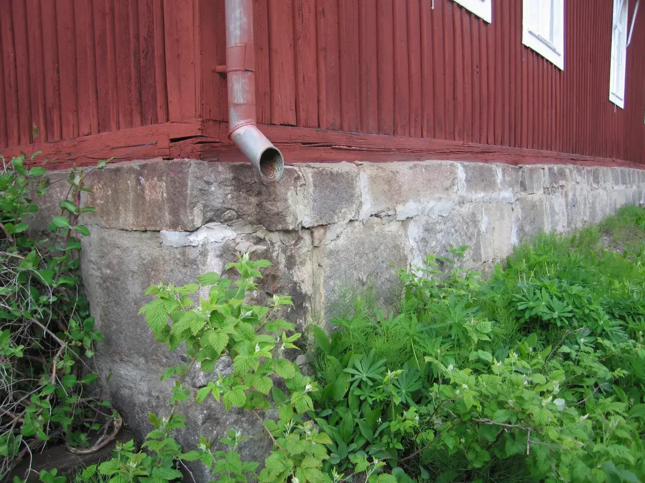 R&ouml;tt tr&auml;hus med stensockel och stupr&ouml;r. Gr&ouml;n v&auml;xtlighet frodas runt husgrunden.