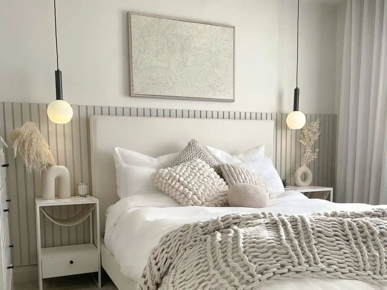 A cozy, small bedroom layout featuring a plush bed with textured pillows and a chunky knit throw. Neutral tones and minimalist decor create a serene atmosphere.