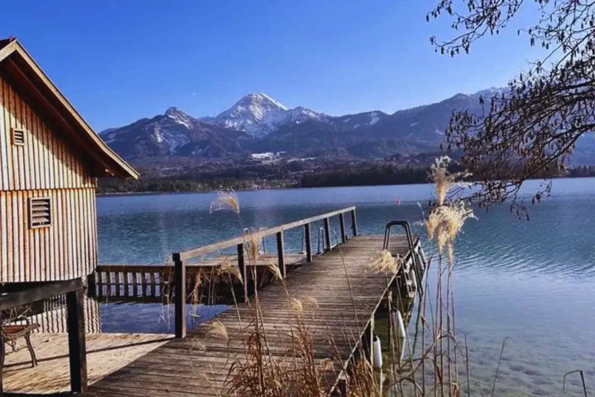 Drewniana chata nad jeziorem w Karyntii, Austria. Drewniany pomost prowadzi do krystalicznie czystej wody, a w tle widać ośnieżone góry.