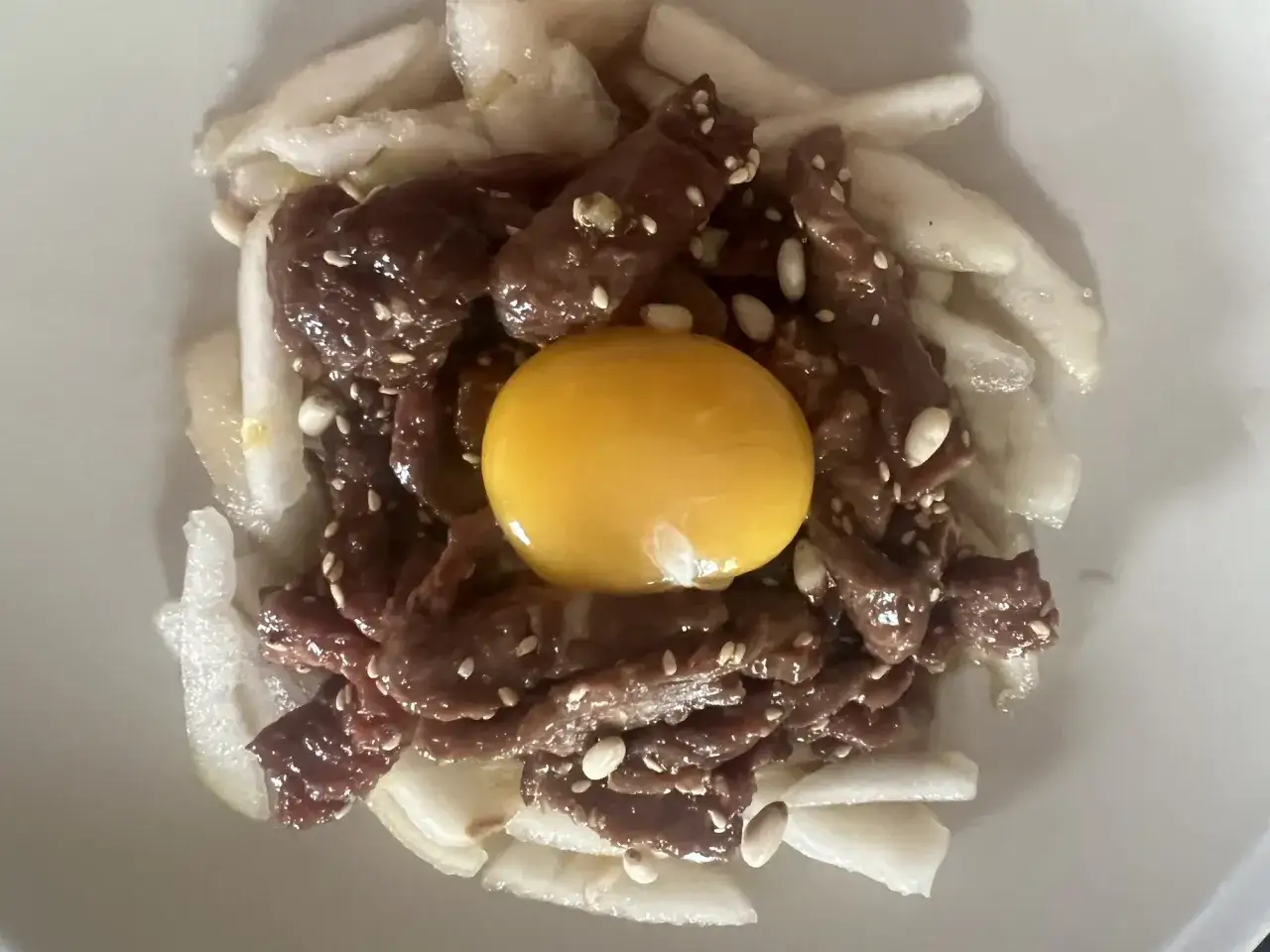 En sk&aring;l med koreansk r&aring;biff, toppad med en gul &auml;ggula, sesamfr&ouml;n och pinjen&ouml;tter, omgiven av tunna skivor av r&auml;ttika.