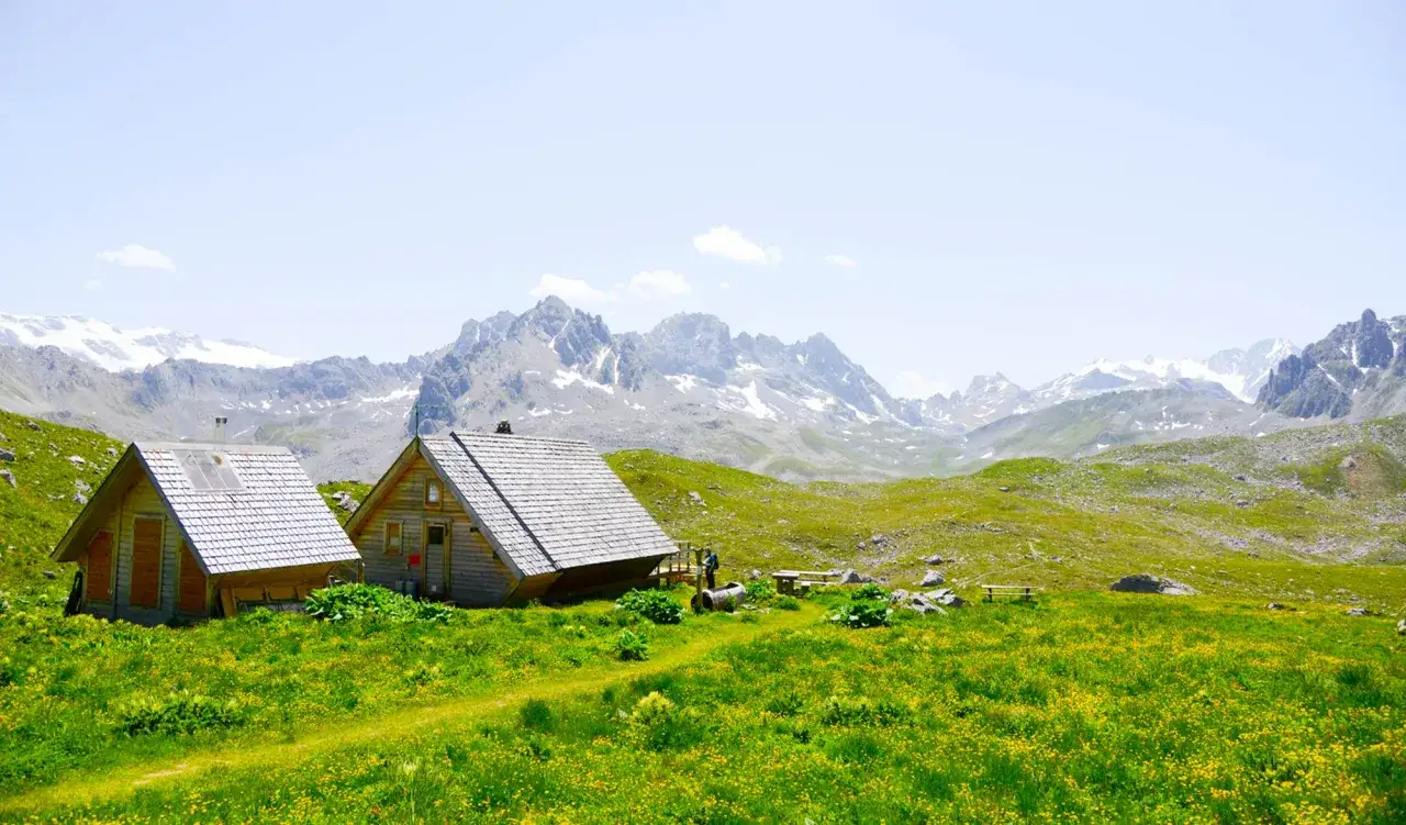 Deux cabanes refuge en bois, nichées dans une prairie fleurie, avec des montagnes enneigées en arrière-plan.