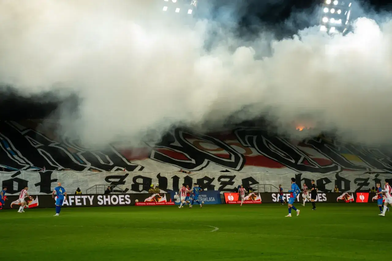 Mecz Cracovia – Piast Gliwice. Gęsty dym zasnuwa trybuny z transparentami, piłkarze biegają po boisku.