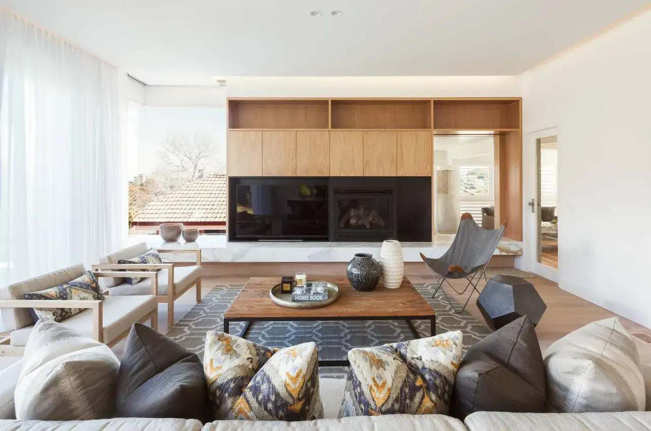 Un elegante sal&oacute;n moderno blanco con sof&aacute;s, chimenea, estanter&iacute;as de madera y una gran ventana.