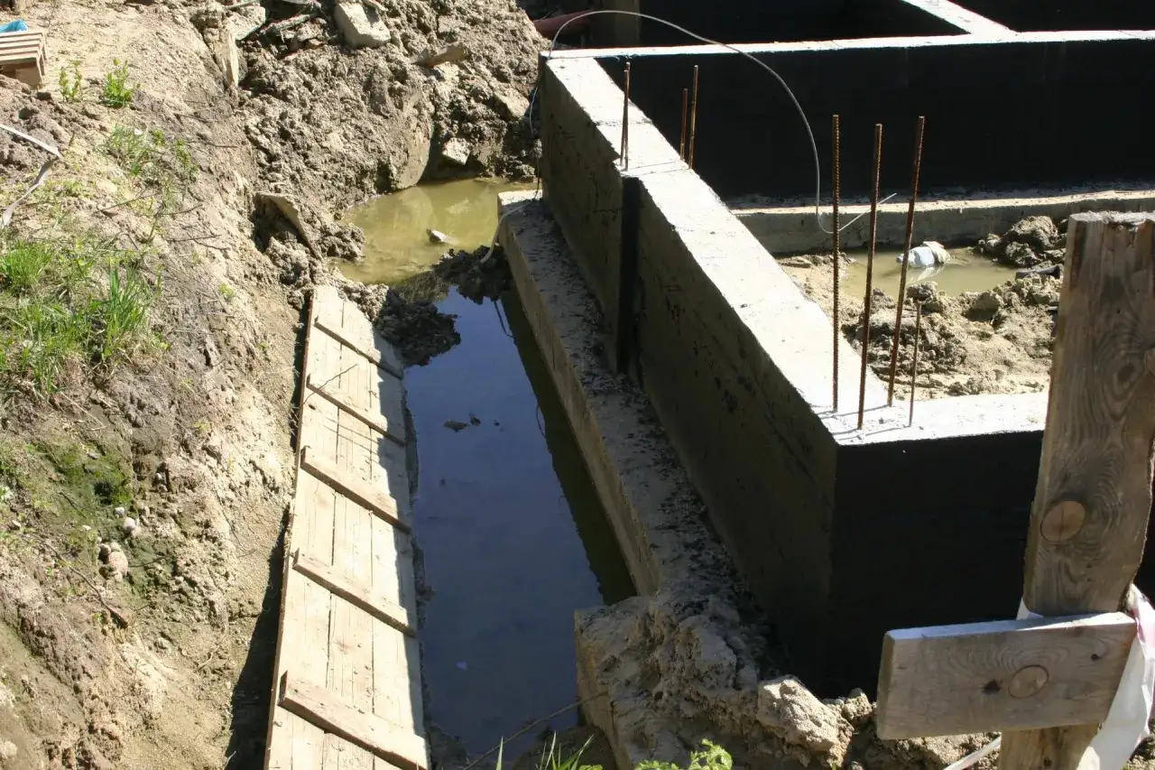 Wykop pod fundamenty wypełniony wodą gruntową, widoczne zbrojenie i beton.
