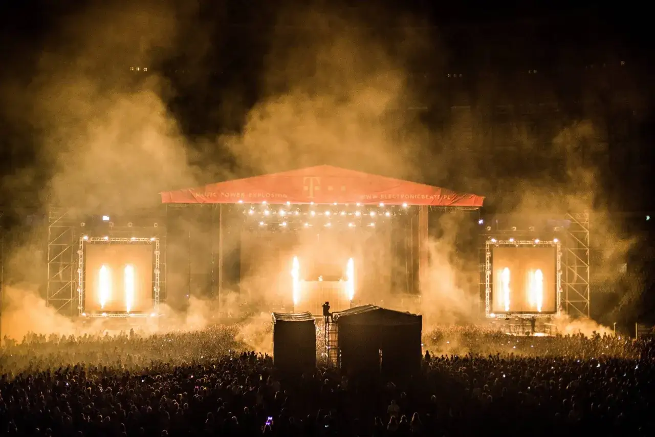 Dorian Tomasiak na scenie festiwalowej, otoczony dymem i światłami. Tłum szaleje pod sceną, a nad nią czerwony baner z logo T-Mobile.