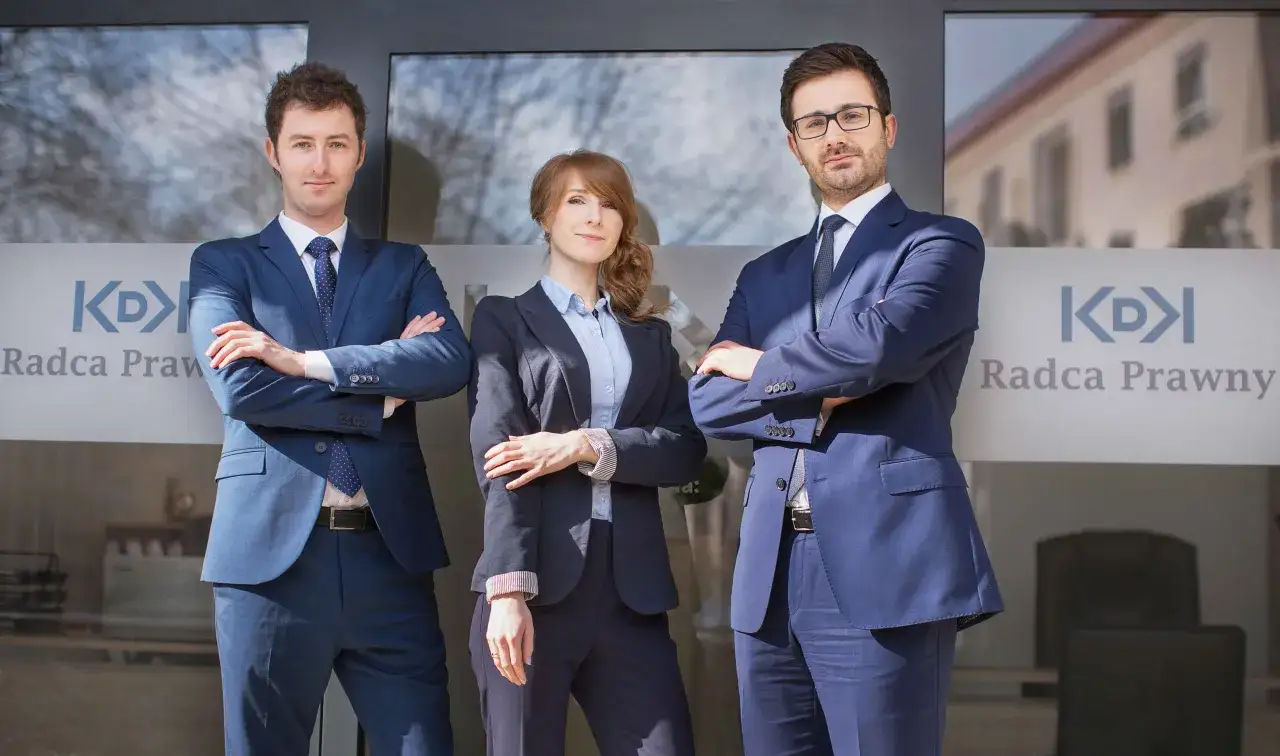 Trzy osoby w garniturach stoją przed budynkiem z napisem