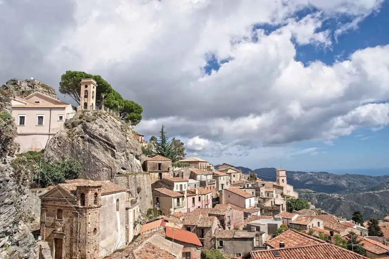 Charming towns in Calabria, Italy, cling to a rocky hillside under a dramatic sky. A bell tower stands sentinel over terracotta roofs.