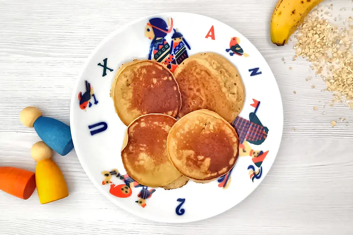 Cuatro tortitas de avena y pl&aacute;tano doradas en un plato infantil con dibujos. Al lado, un pl&aacute;tano y copos de avena.