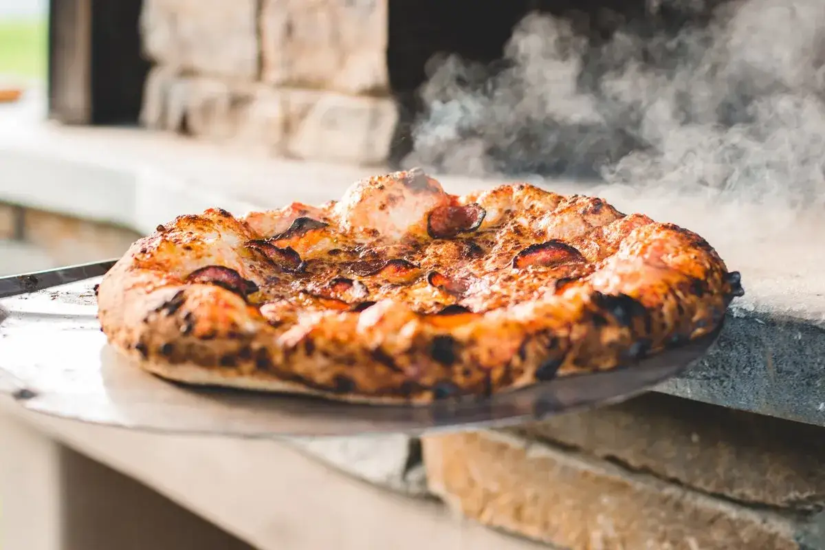 Gorąca pizza z pepperoni na metalowej łopatce, gotowa do przełożenia na kamień. Dym unosi się znad wypieku.