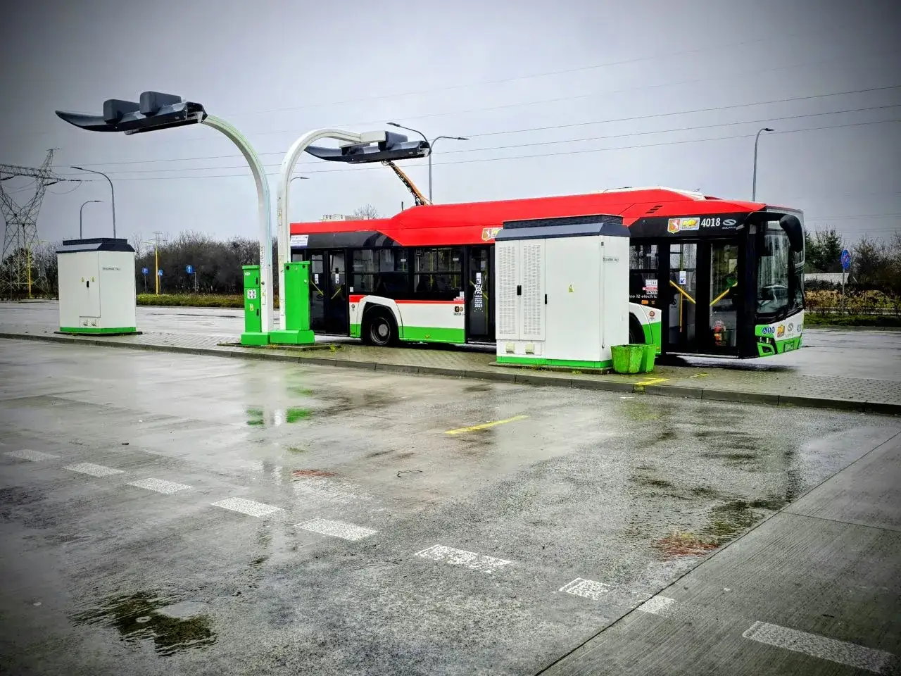 Elektryczny autobus czeka na odjazd. Na deszczowym placu ładowania, gdzie widoczny jest 43 rozkład jazdy, stoi nowoczesny pojazd.