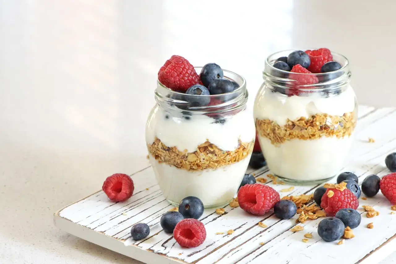 Due vasetti di yogurt a colazione con granola, lamponi e mirtilli freschi, su un tagliere di legno bianco.