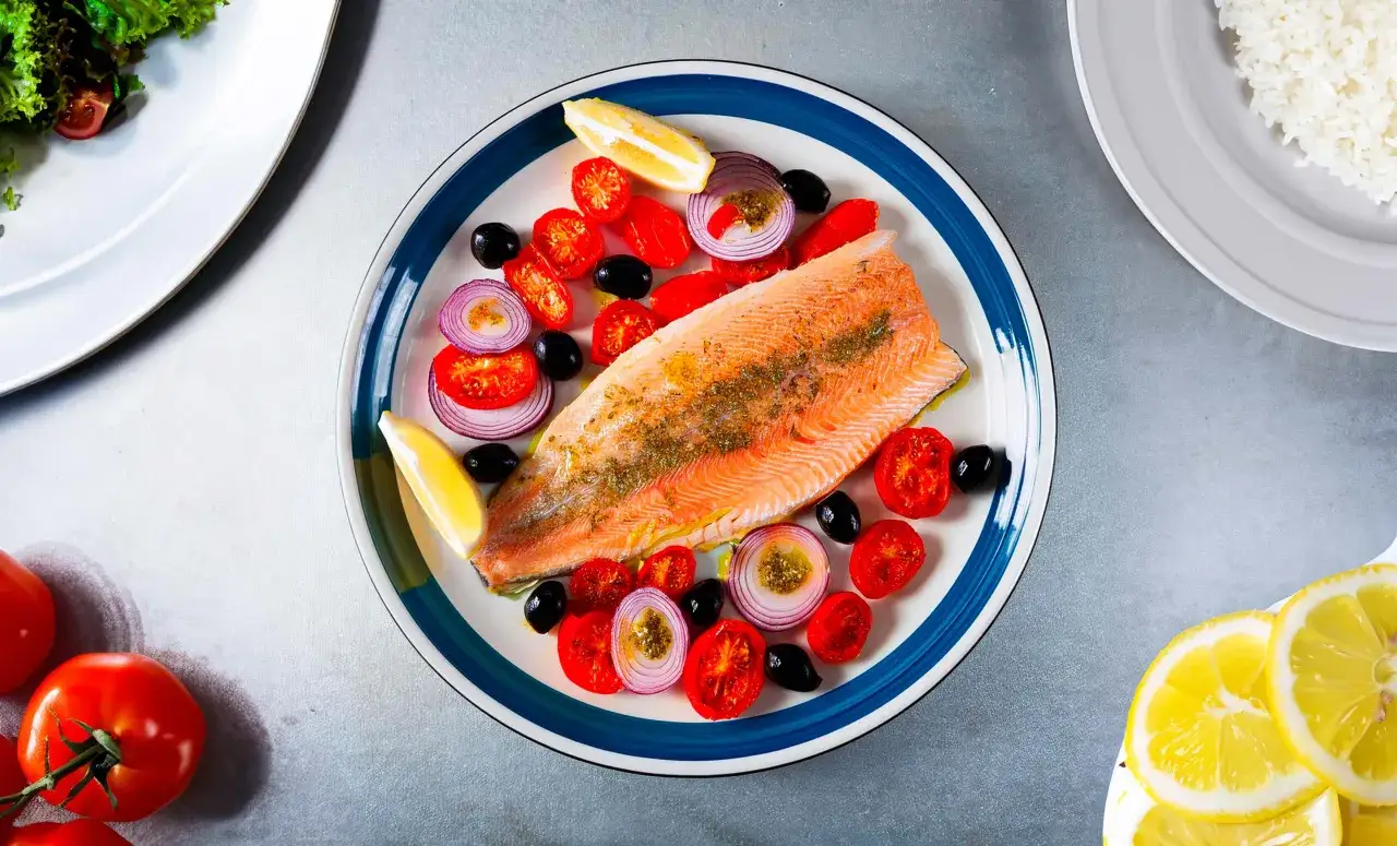 Une assiette de truite au four, garnie de tomates cerises, d'oignons rouges, d'olives noires et de quartiers de citron.