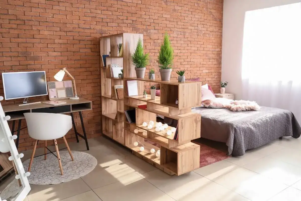 Un ambiente moderno con una **cucina e camera da letto insieme**, separate da una libreria in legno. Parete in mattoni a vista.