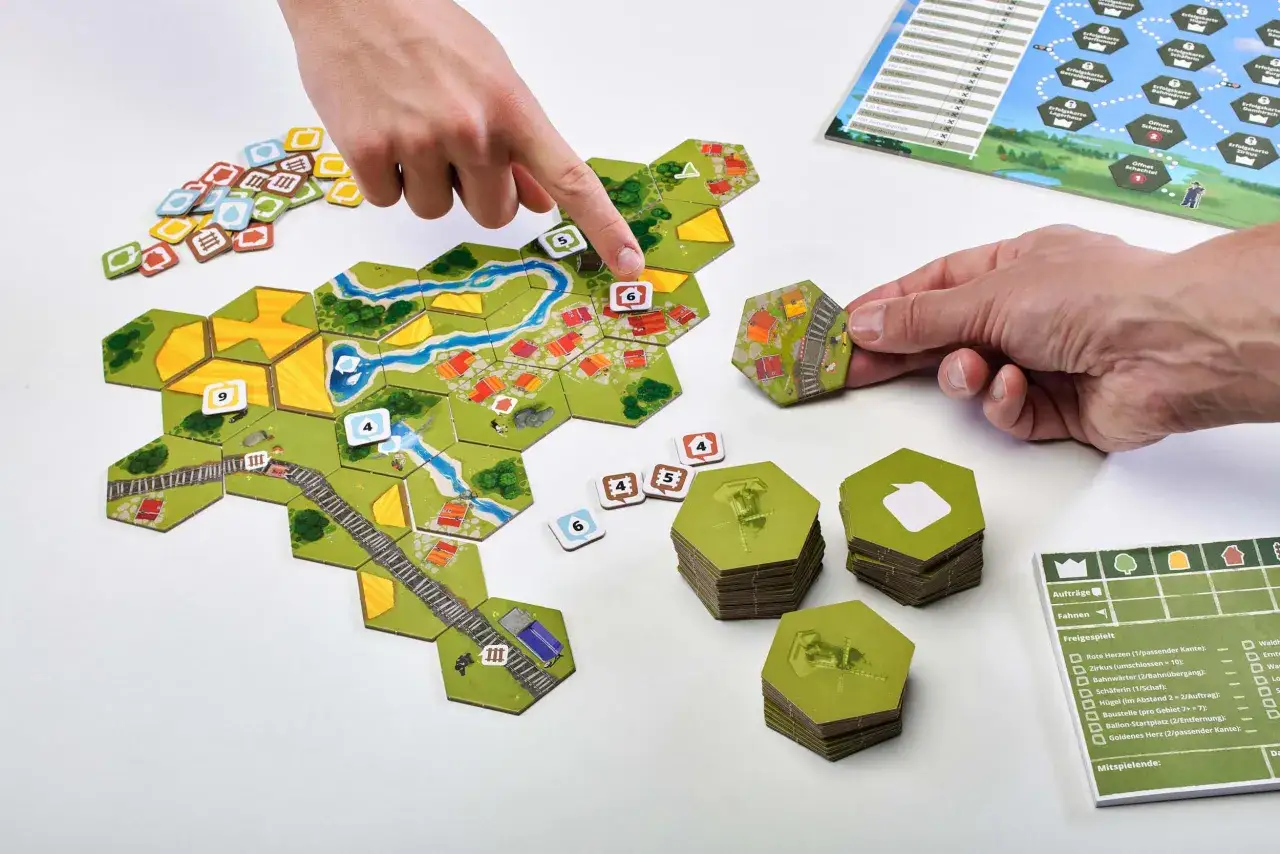 H&auml;nde legen Spielsteine f&uuml;r das Dorfromantik Spiel des Jahres an. Ein Spieler platziert eine Bahnsteig-Kachel, w&auml;hrend andere Spielsteine und das Spielbrett zu sehen sind.