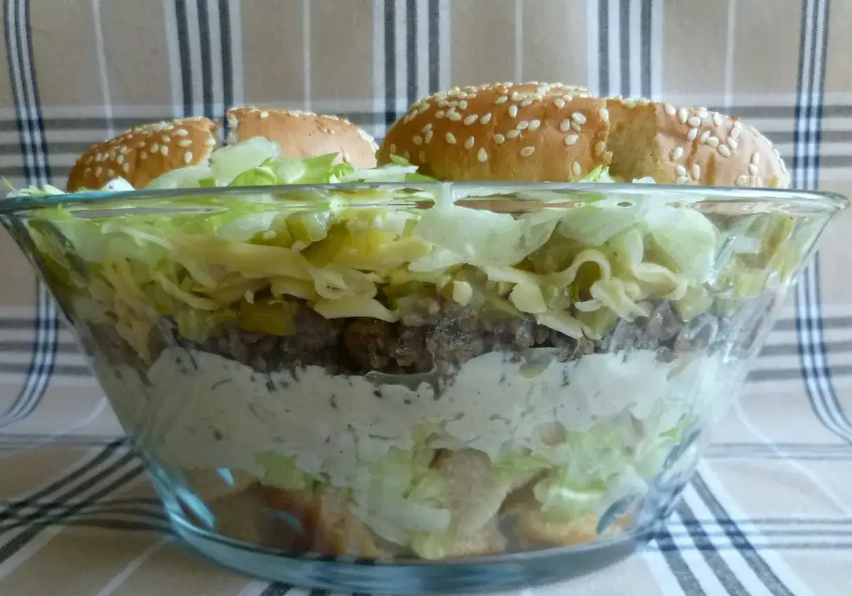 Warstwowa sałatka big mac przepis: mięso mielone, ser, sałata, ogórki i sos. Na wierzchu bułki sezamowe.