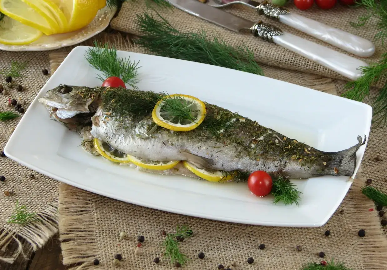 Pstrąg z piekarnika, pieczony z cytryną i ziołami, podany na białej tacy z pomidorkami koktajlowymi i koperkiem.