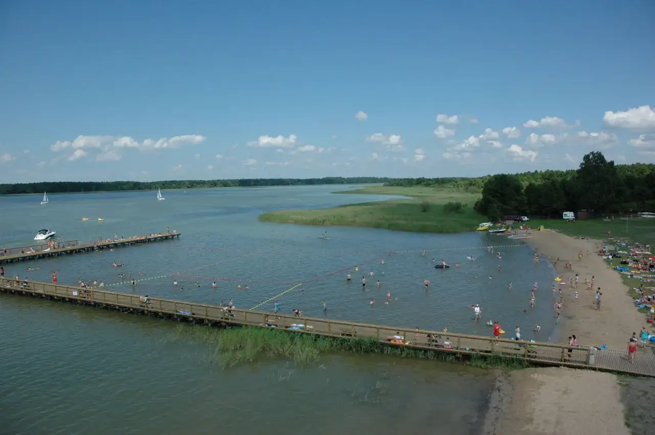 Letnie popołudnie w mieście nad jeziorem. Ludzie kąpią się, opalają na plaży i spacerują po drewnianych pomostach.