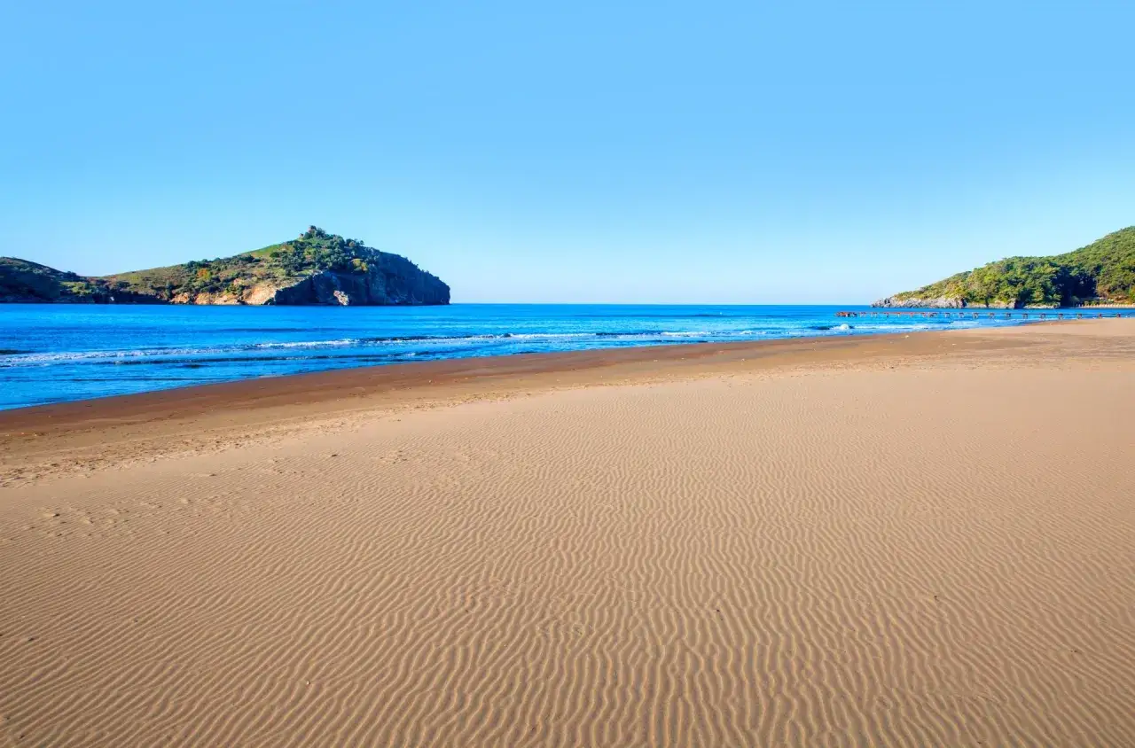 Piękna plaża w Sarigerme, z piaszczystym wybrzeżem i turkusowym morzem. W oddali widać zielone wzgórza i skaliste wysepki.