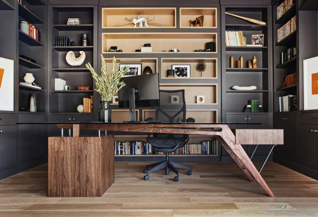 A modern office with dark grey built-in cabinets and open shelving displaying books and decor. A unique wooden desk with a glass top sits center, complemented by an ergonomic chair.