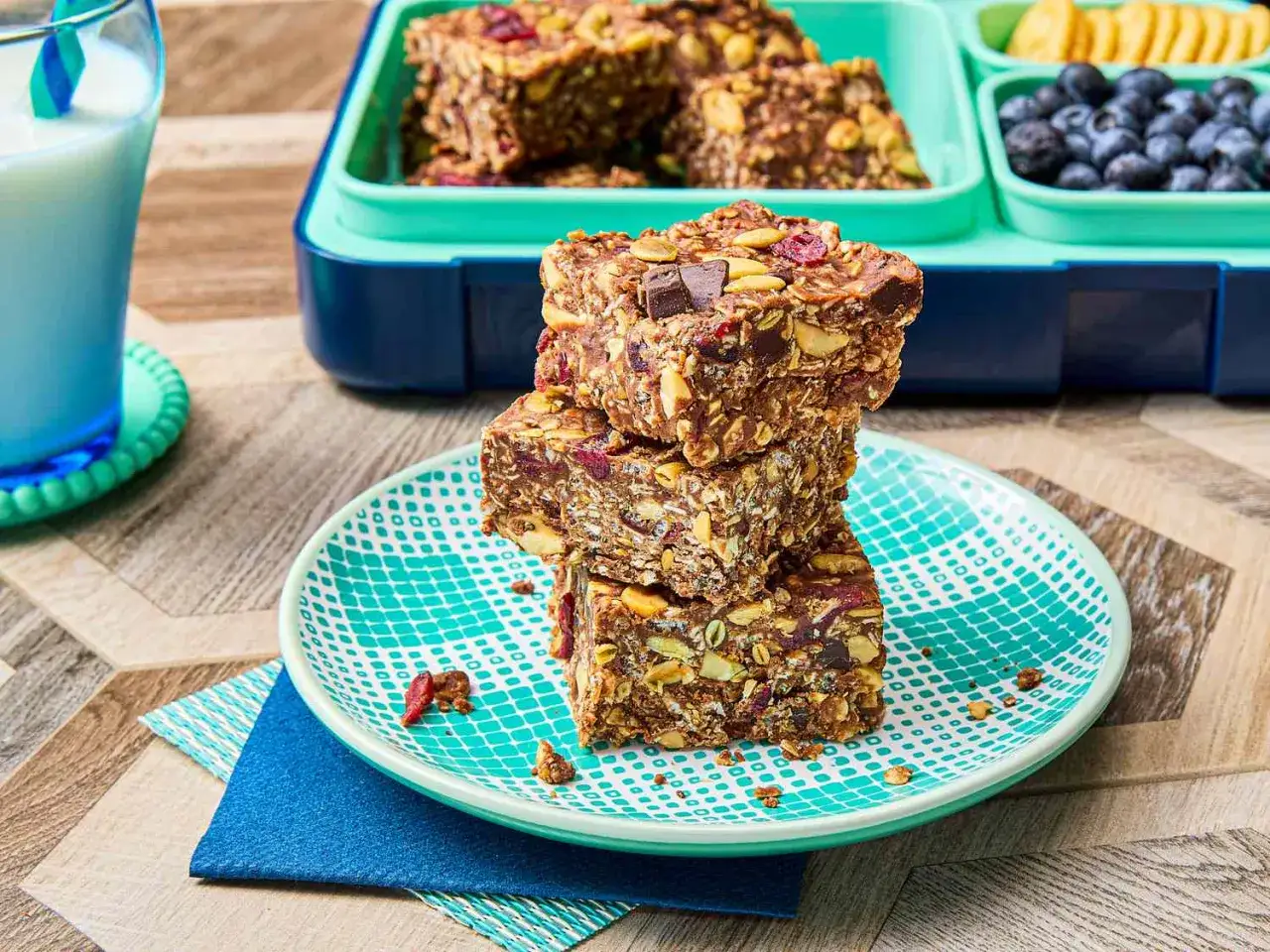 H&auml;r &auml;r ett enkelt proteinbars recept: staplade bars med n&ouml;tter, torkad frukt och choklad p&aring; en tallrik. En matl&aring;da med bl&aring;b&auml;r och kex syns i bakgrunden.