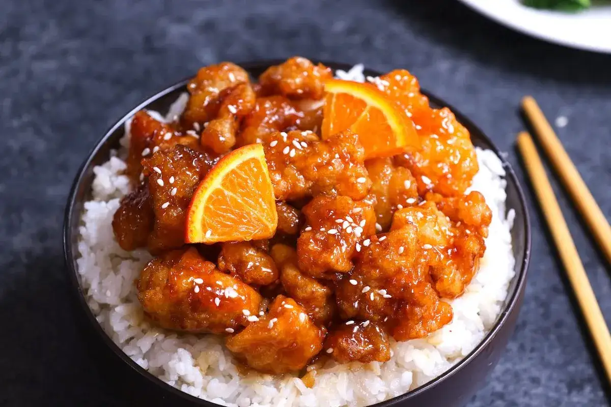Skål med fluffigt ris toppad med glaserad orange chicken, garnerad med apelsinskivor och sesamfrön.