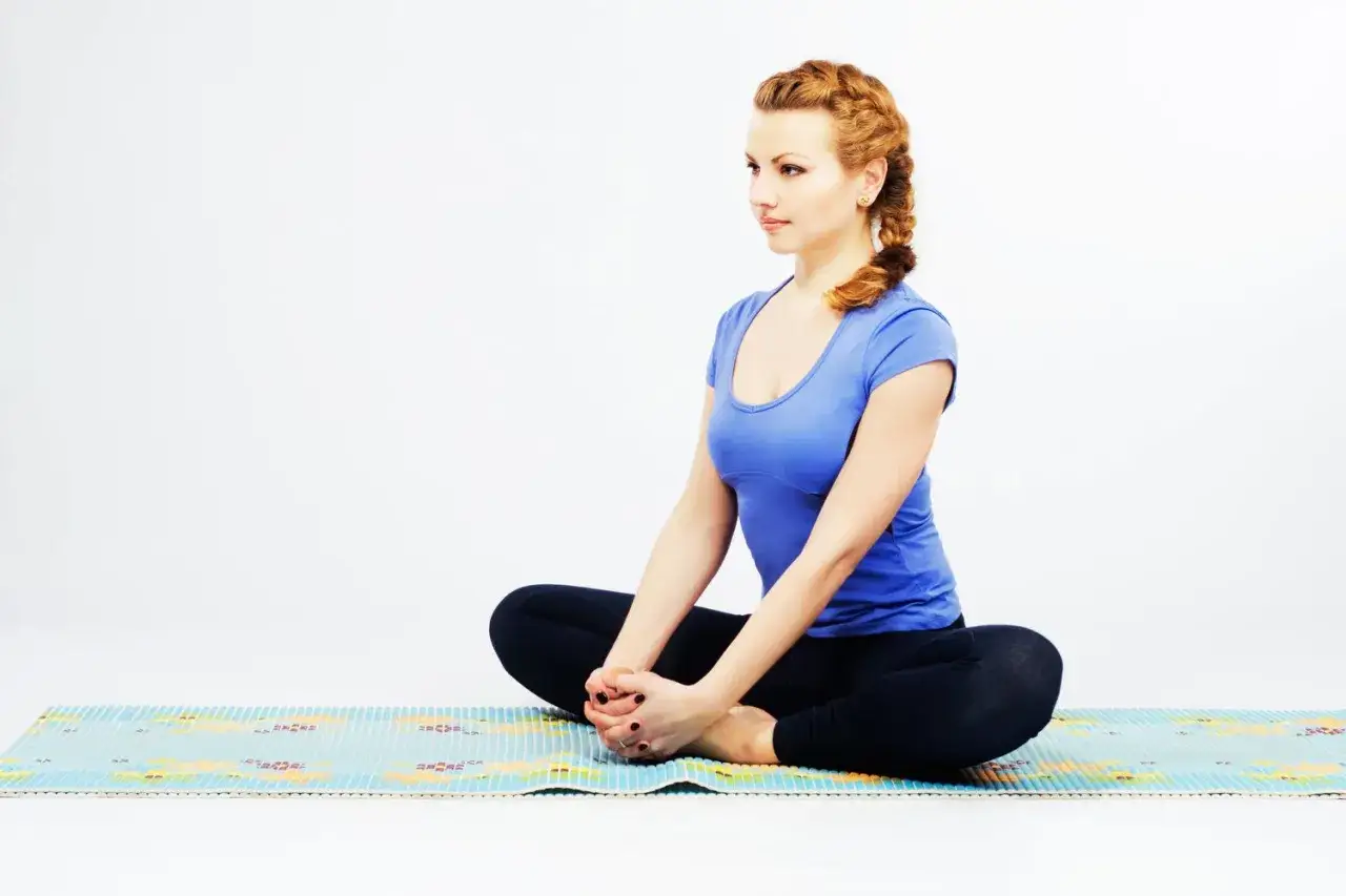 Frau in blauem Shirt und Leggings sitzt auf Yogamatte, Beine im Schneidersitz, um die Leiste zu dehnen.