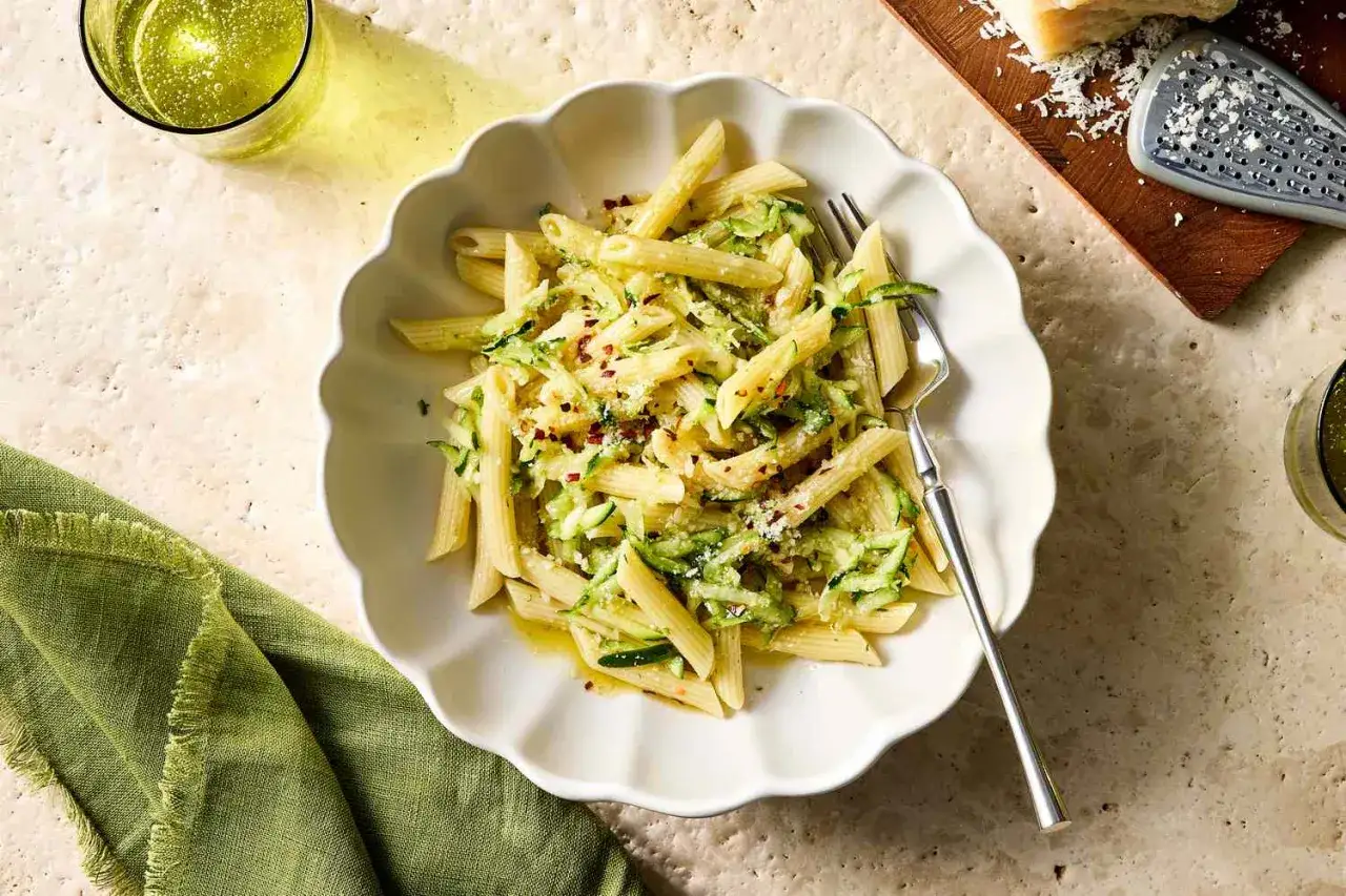 Penne z cukinią, posypane parmezanem i płatkami chili, podane w białej misce z zieloną serwetką.