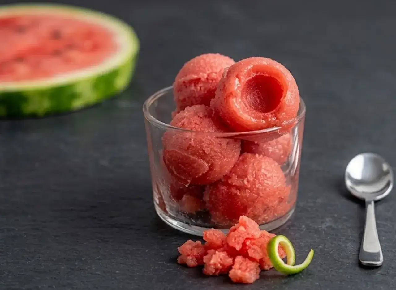 Sorbetto all'anguria senza gelatiera in un bicchiere, con una fetta di anguria sfocata sullo sfondo e una scorza di lime.