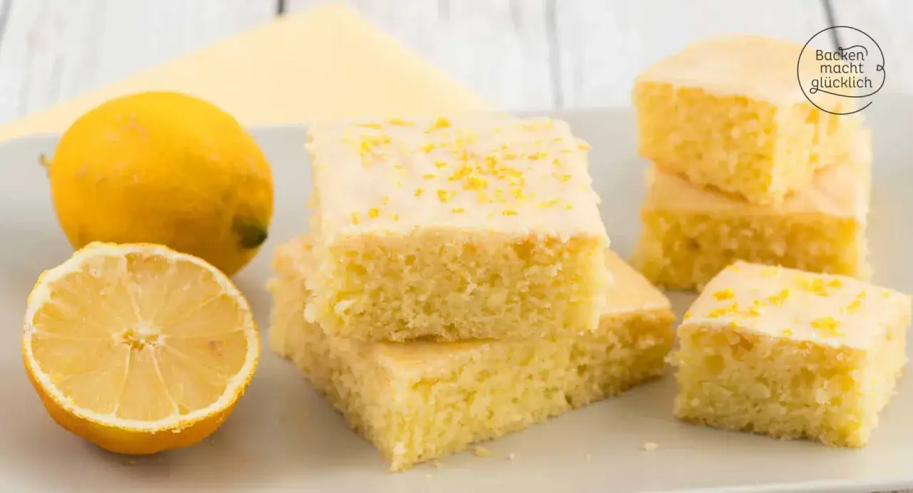Saftiger Zitronenkuchen vom Blech, bestreut mit Zitronenschale. Daneben liegen frische Zitronen.