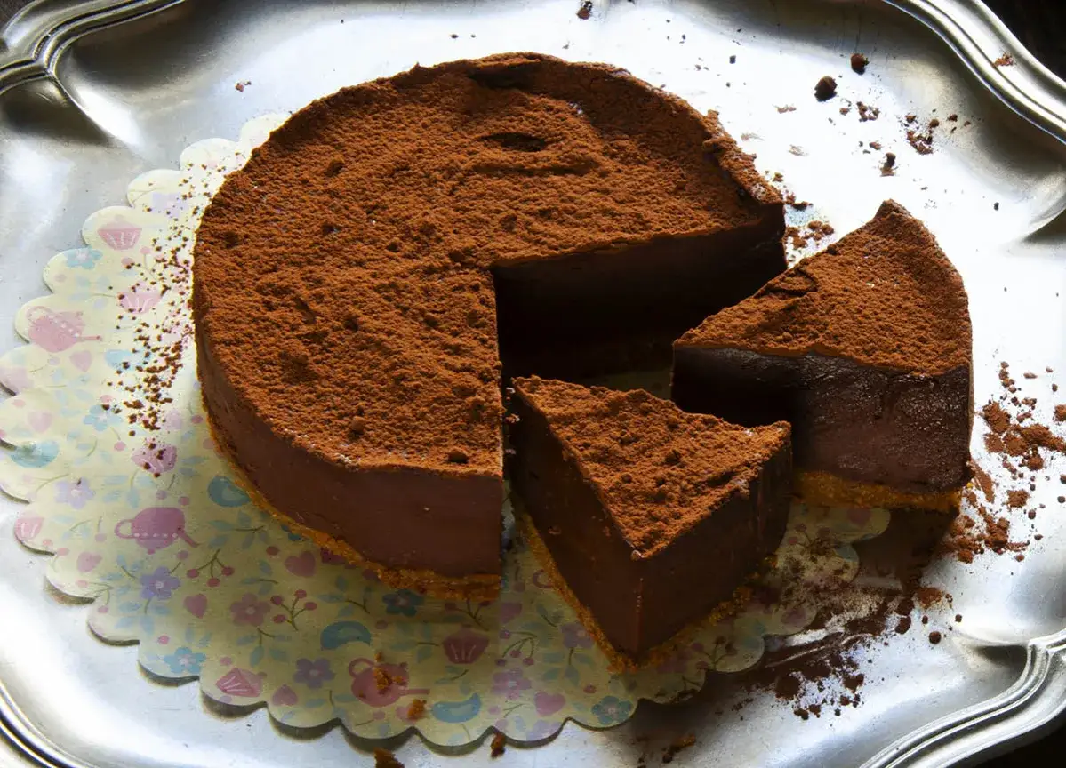 Deliciosa marquesa de chocolate, cubierta de cacao en polvo, servida en una bandeja plateada sobre un papel decorado con flores.