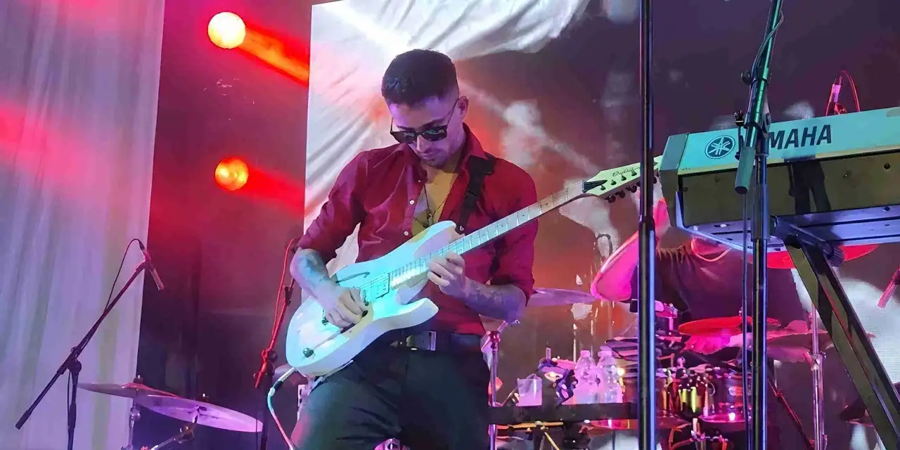 Mężczyzna w czerwonej koszuli gra na gitarze, jak jeden z najszybszych gitarzystów świata. Na scenie światła i instrumenty.