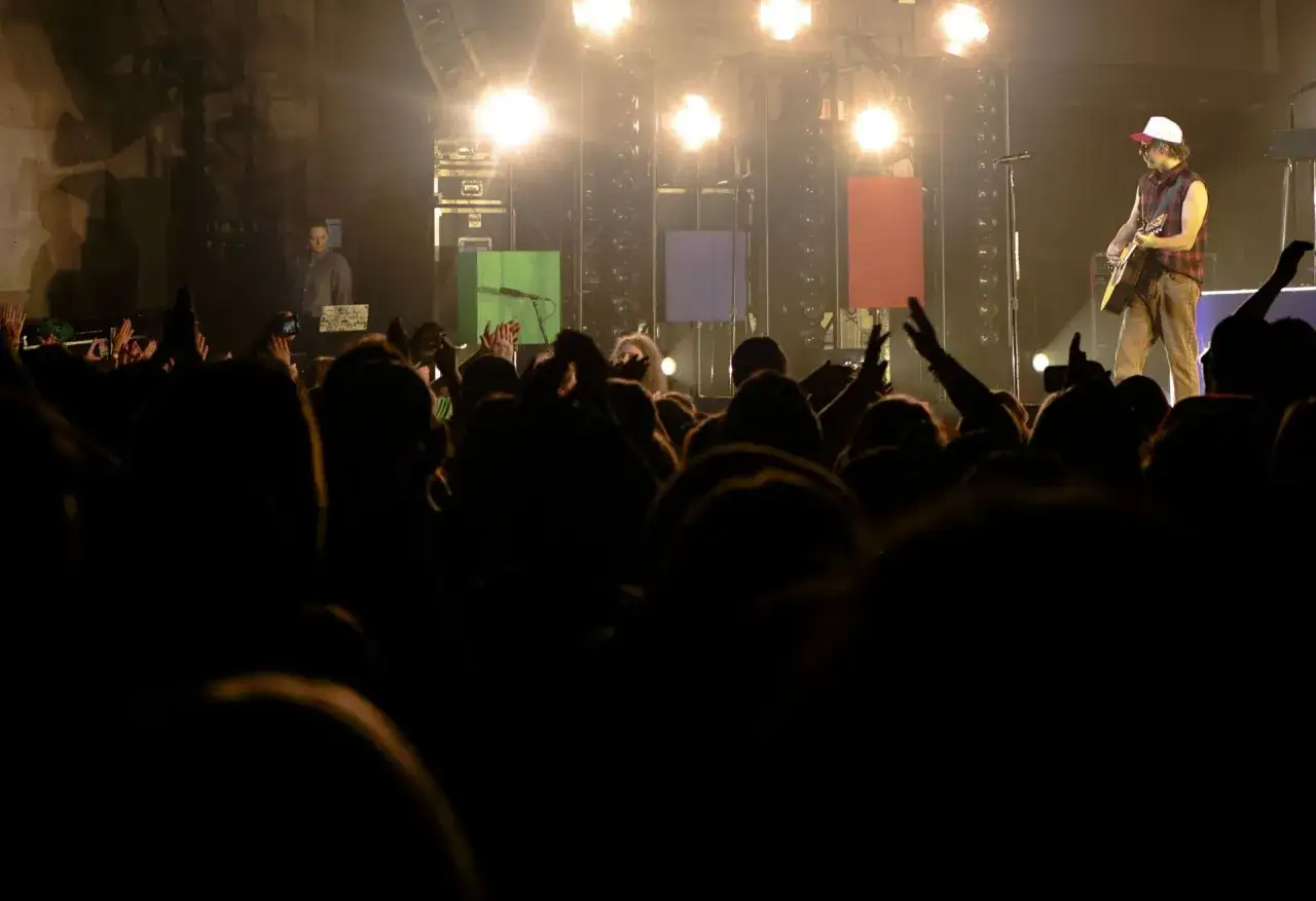 Koncertowy tłum szaleje przy dźwiękach punk rocka. Na scenie gitarzysta w kratę i czapce, za nim kolorowe panele i światła.
