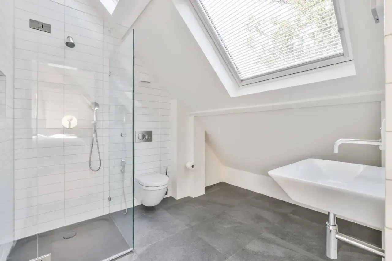 Moderno baño en buhardilla con ducha de cristal, inodoro y lavabo bajo una ventana inclinada.