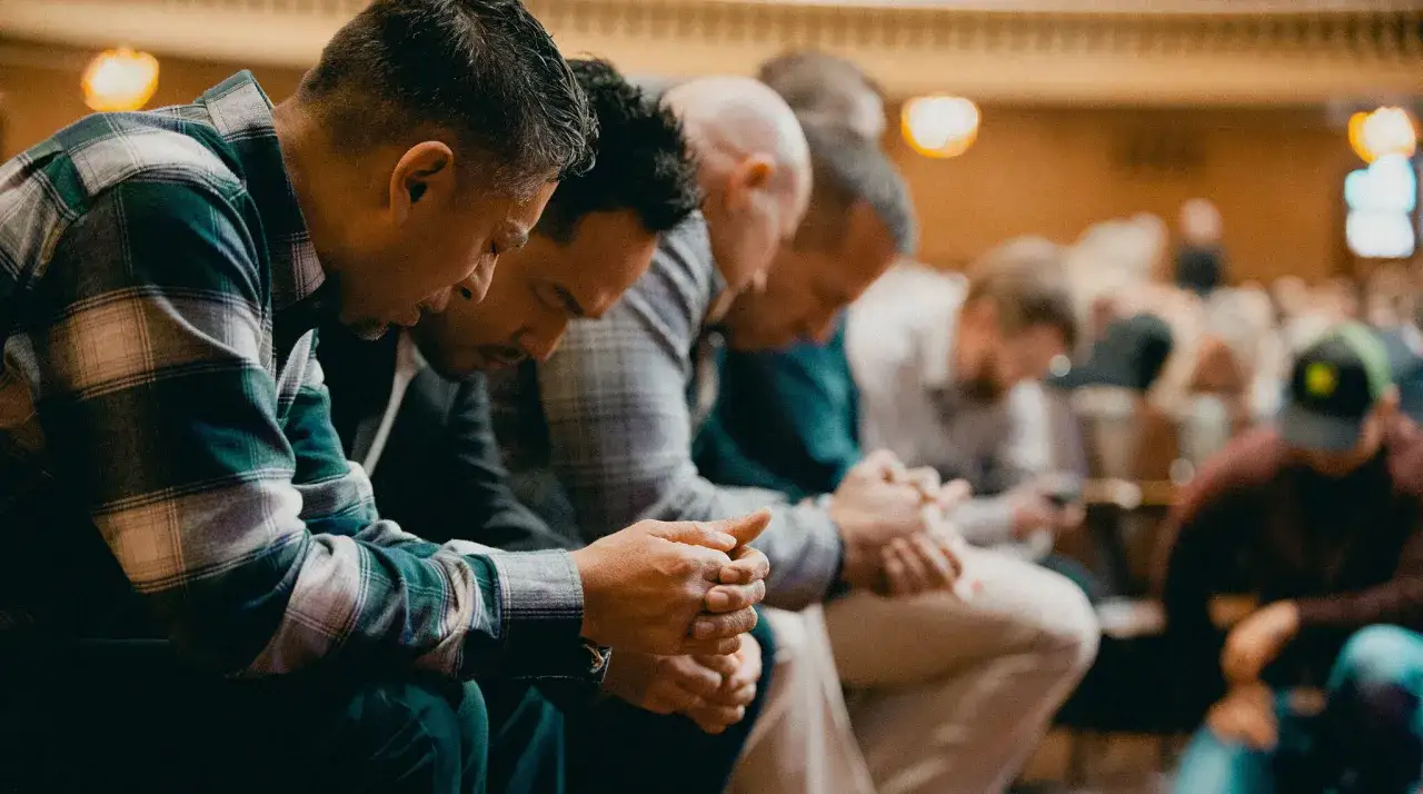 Men in a row, heads bowed in prayer. Their hands are clasped, a moment of quiet reflection. Prayers requested.