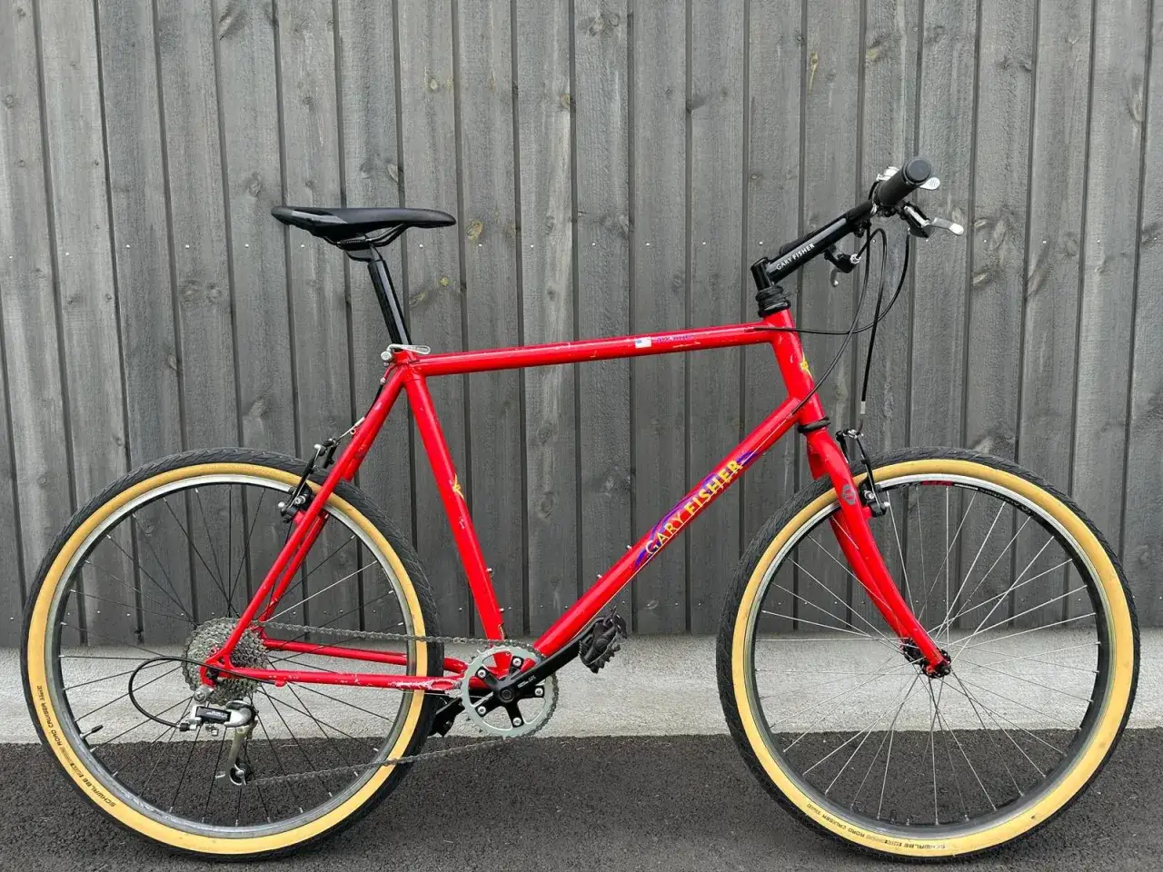 A vintage red Gary Fisher Advance mountain bike with tan tires stands against a wooden fence.