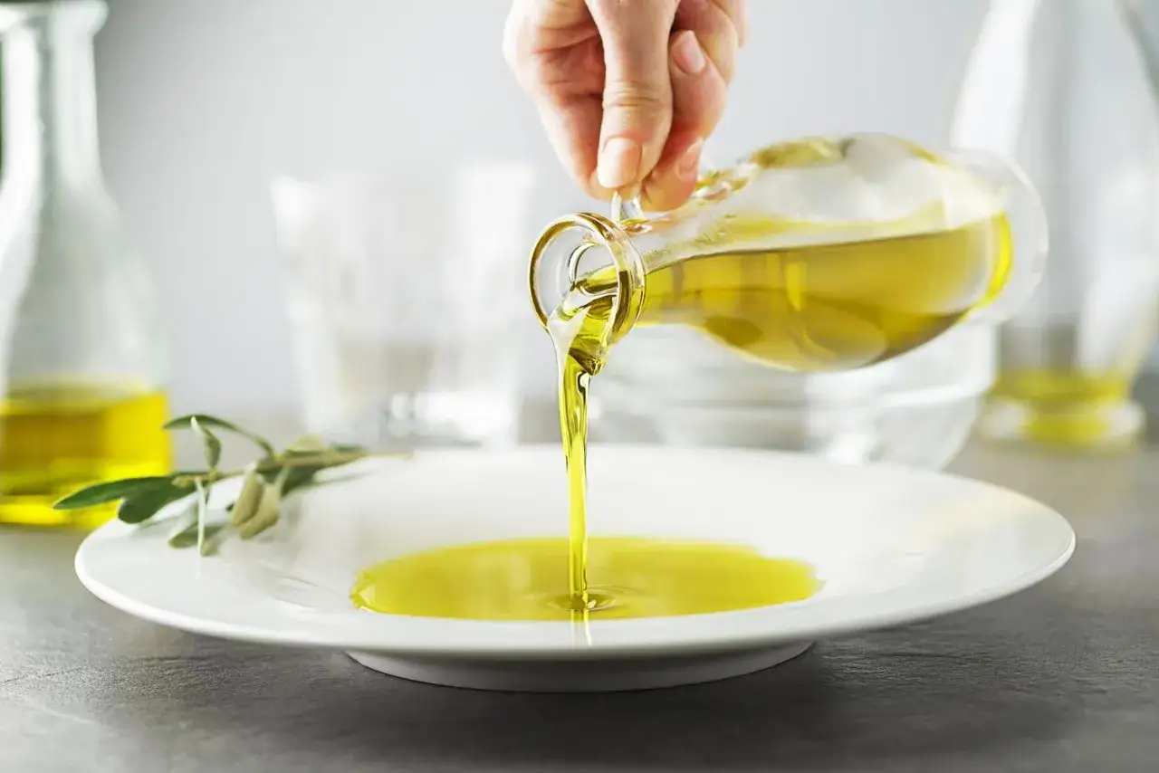 Una mano vierte aceite de oliva en un plato blanco. Este aceite de oliva tiene vitamina D, un nutriente esencial.