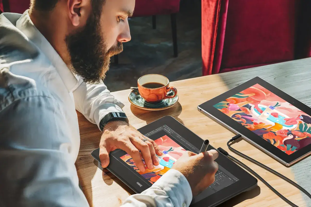 Mężczyzna z brodą tworzy cyfrowy obraz na tablecie graficznym, używając pi&oacute;rka. Obok stoi filiżanka kawy i drugi tablet z kolorową grafiką.