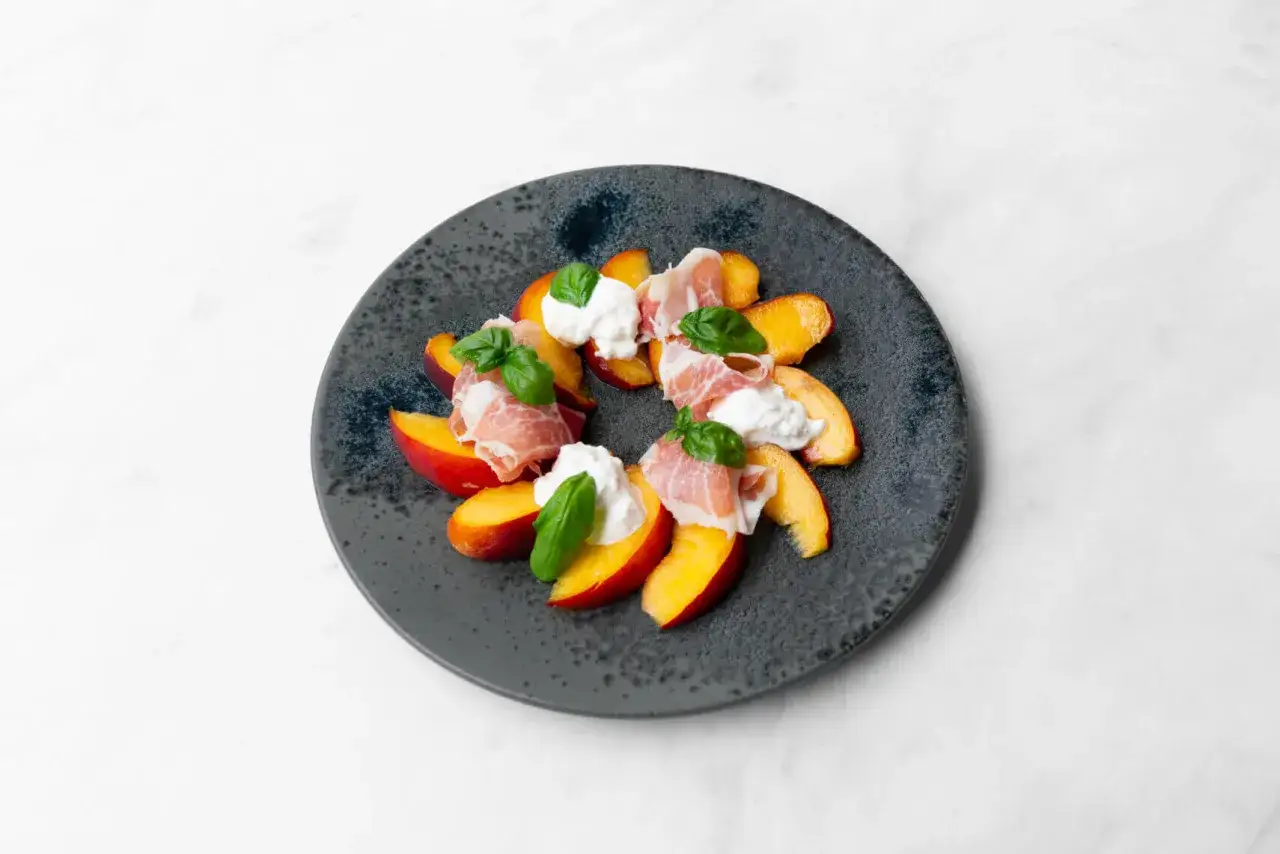 Ein Teller mit Pfirsichspalten, belegt mit Burrata, Prosciutto und Basilikum. Ein einfaches Gericht, das zeigt, wie vielseitig Burrata-Rezepte sein können.