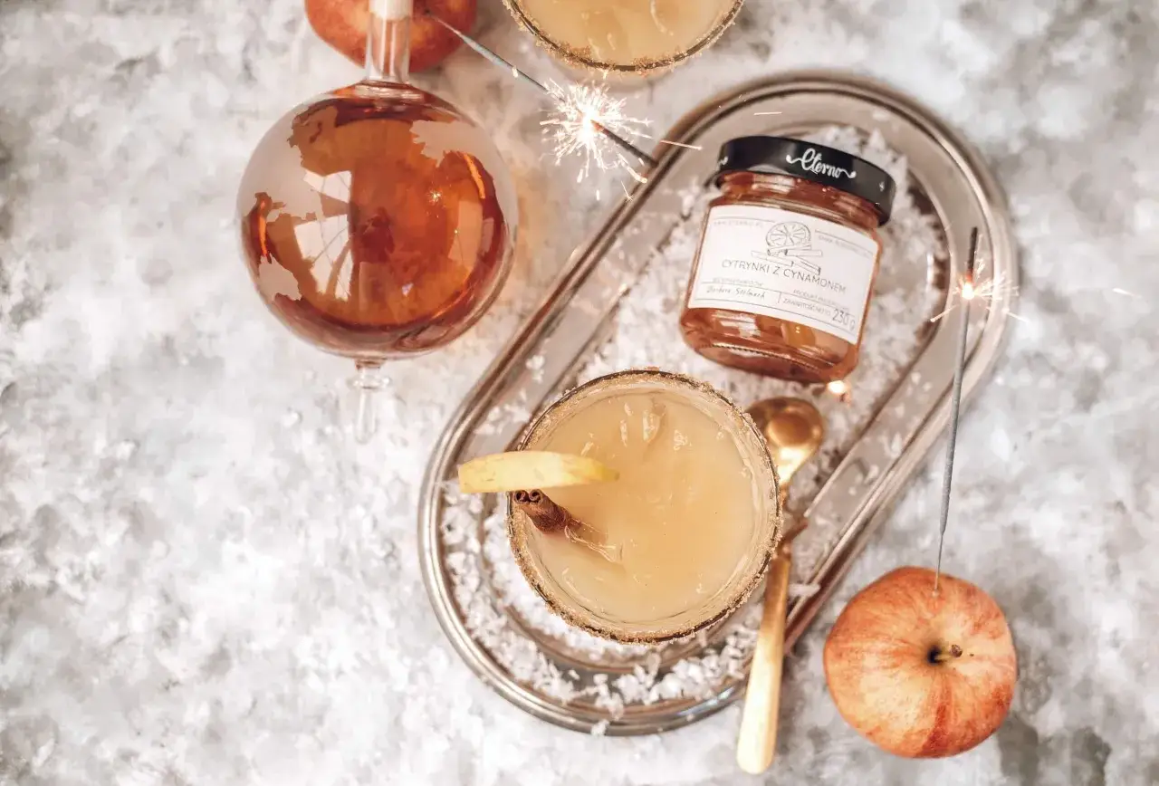 Elegancki drink: whisky z sokiem jabłkowym, ozdobiony laską cynamonu i plasterkiem jabłka. Na tacy błyszczą zimne ognie, a obok stoi słoiczek z cytrynkami i cynamonem.