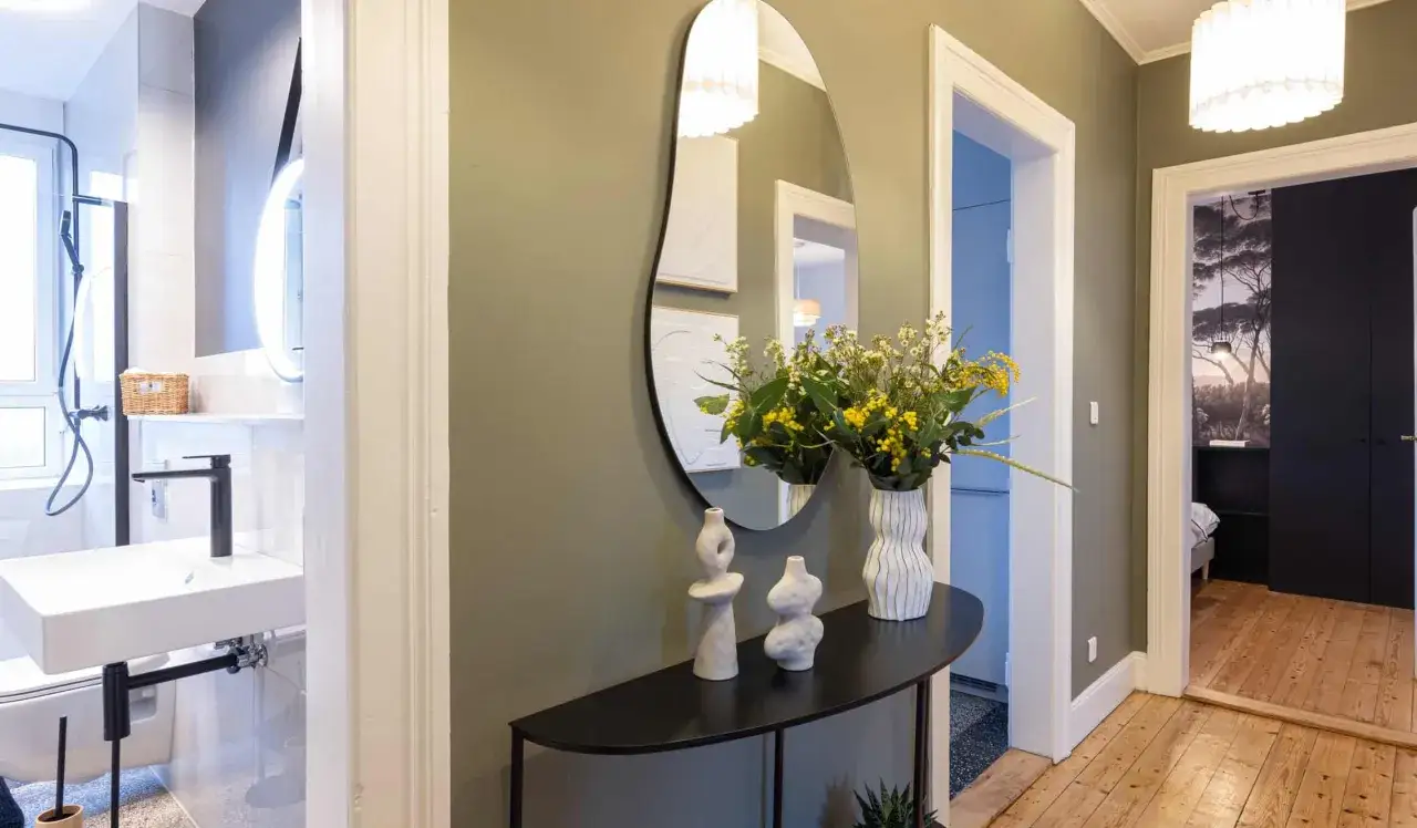 Un couloir de maison accueillant avec un miroir organique, des vases d&eacute;coratifs, un bouquet de fleurs jaunes et une vue sur une salle de bain et une chambre.