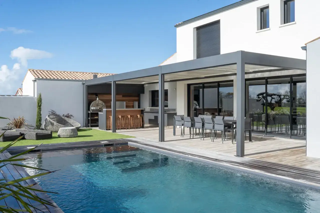 Un &eacute;l&eacute;gant pool house avec cuisine d'&eacute;t&eacute;, bar et coin repas, donnant sur une piscine bleue.