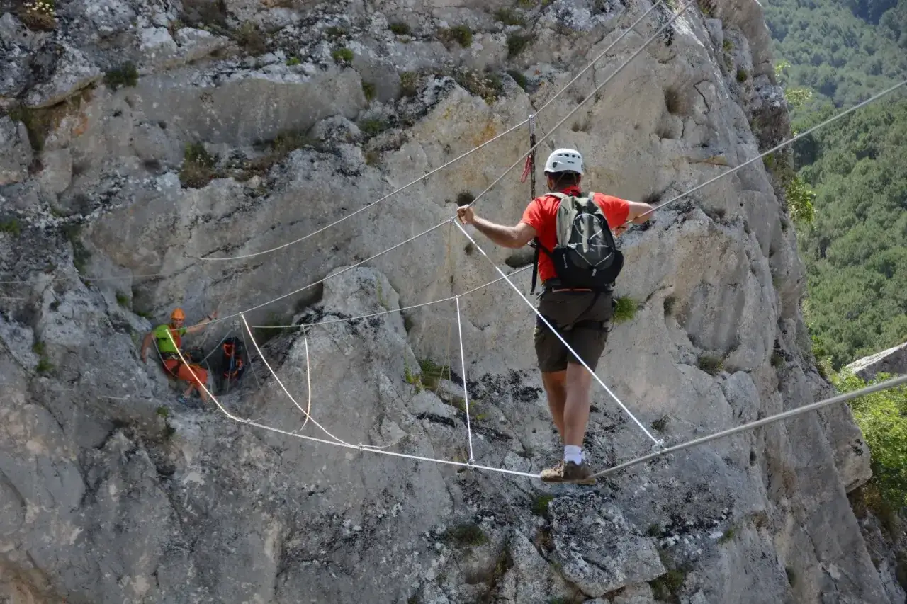 Hombre con casco y mochila cruza un puente colgante, parte del material de vía ferrata, sobre un barranco rocoso.