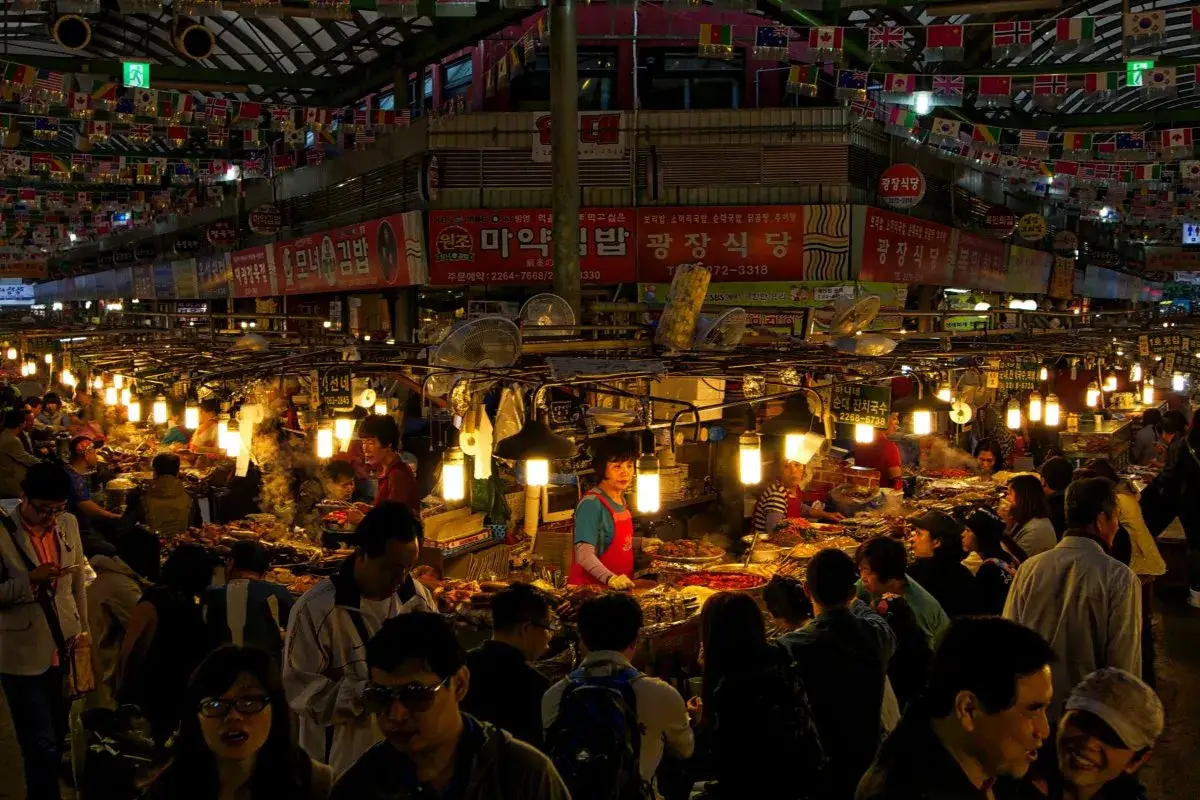 Livligt Gwangjang Market i Seoul, med matst&aring;nd, folk och flaggor som h&auml;nger fr&aring;n taket.