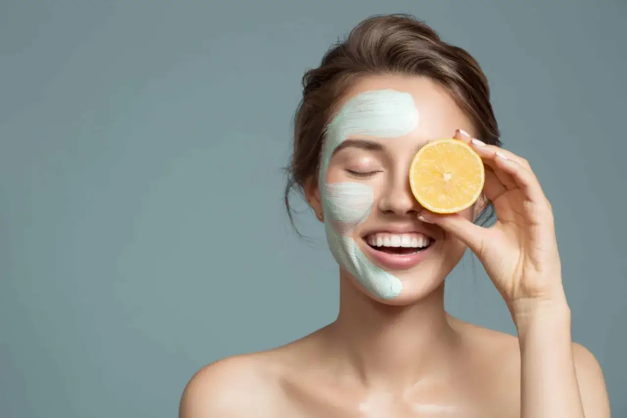 Une femme souriante avec un masque facial vert clair et une tranche de citron devant son &oelig;il, illustrant les bienfaits du citron sur la peau.