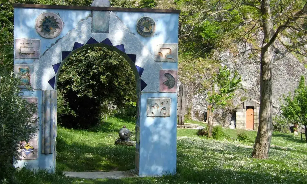 Arco decorato con mosaici e ceramiche, un invito a scoprire cosa vedere a Pennabilli. Un piccolo edificio in pietra e un giardino fiorito completano la scena.