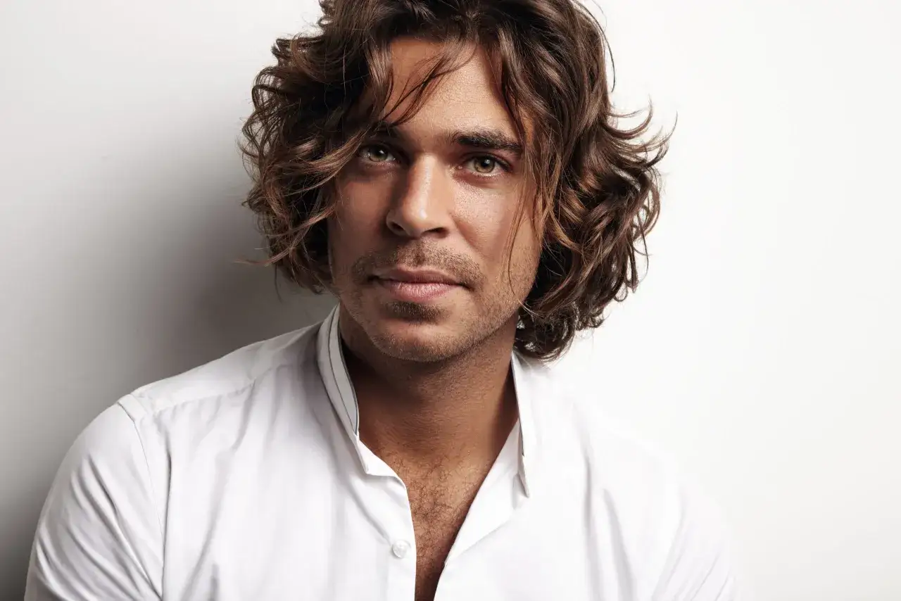 Hombre con pelo castaño ondulado, un estilo que inspira sobre como dejarse media melena hombre. Viste camisa blanca.