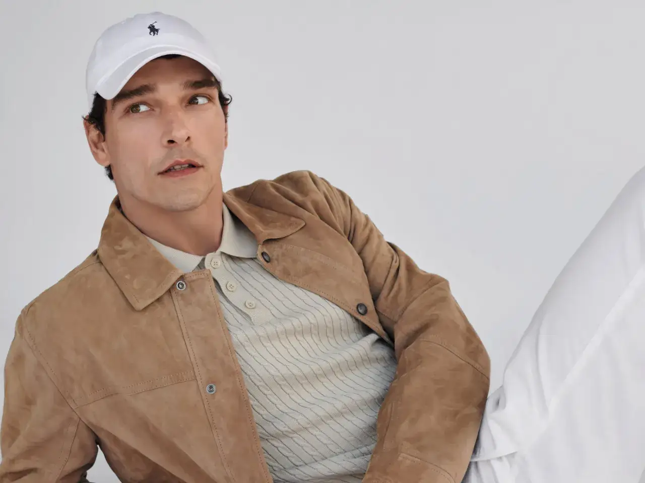 Mann mit brauner Wildlederjacke, beigem Poloshirt und wei&szlig;er Kappe mit Polo-Logo.