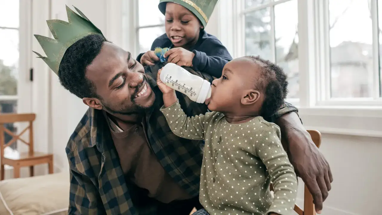 Ein l&auml;chelnder Papa mit gr&uuml;ner Krone f&uuml;ttert sein Baby mit der Flasche, w&auml;hrend sein &auml;lterer Sohn daneben spielt. Tolle Eigenschaften eines Papas!