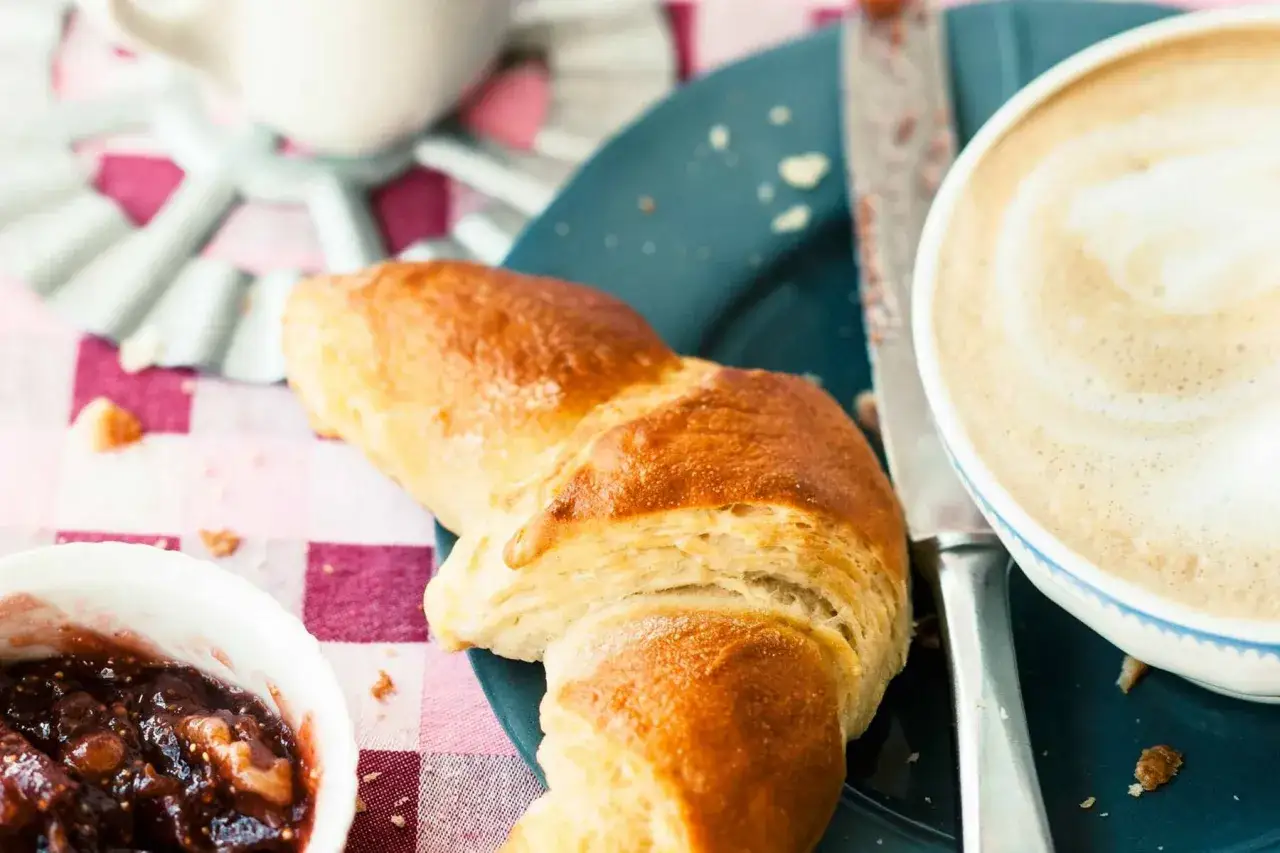 Ein goldbraunes Croissant neben einer Tasse Kaffee au lait und Feigenmarmelade auf kariertem Stoff.