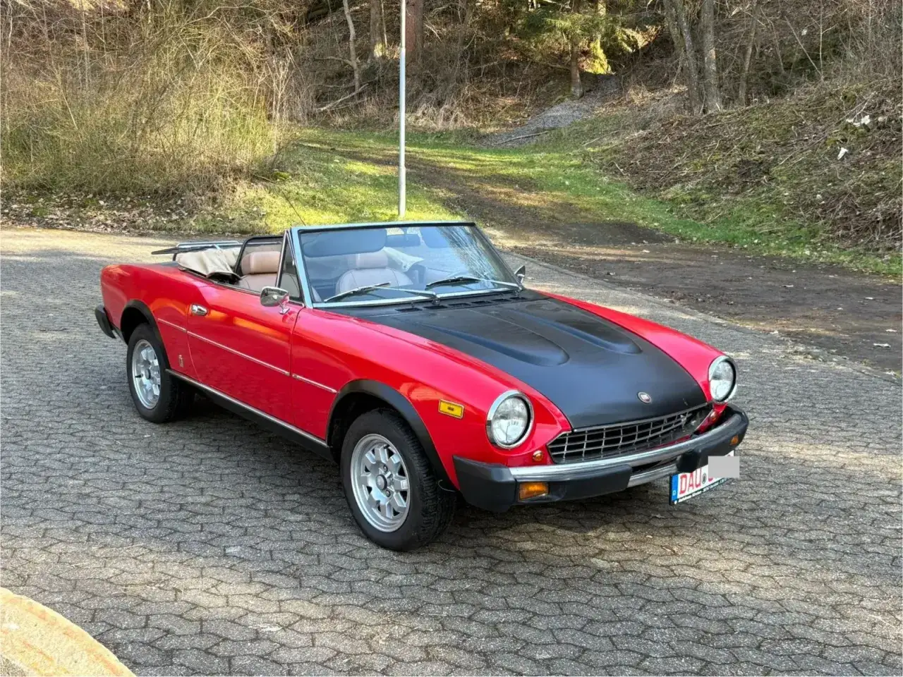 Czerwony fiat 124 spider z czarną maską stoi na brukowanej drodze, otoczony zielenią i drzewami.